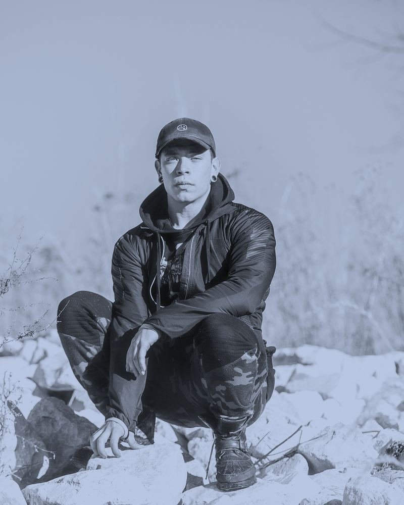 Jason Osaj crouched on rocky ground in a hooded jacket and cap, looking directly at the camera